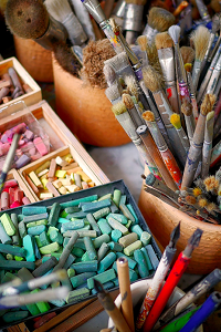 A collection of coloured chalk and pots full of paint brushes.