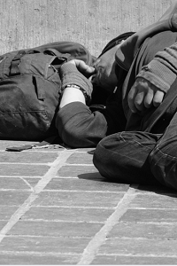 A black and white picture of a man sleeping on the street with a backpack as a pillow.