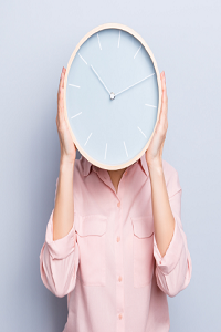 Person holding large clock face in front of their head, they are wearing a salmon coloured shirt