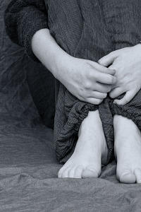 A black and white picture of a young person sitting barefoot with their hands reached around their knees which have been pulled up to their chest.