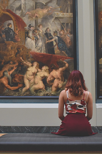 A woman sits in front of a collection of classical paintings in a museum.