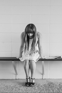 A black and white picture of a young girl in a dress sitting on the bench looking downwards with worry.