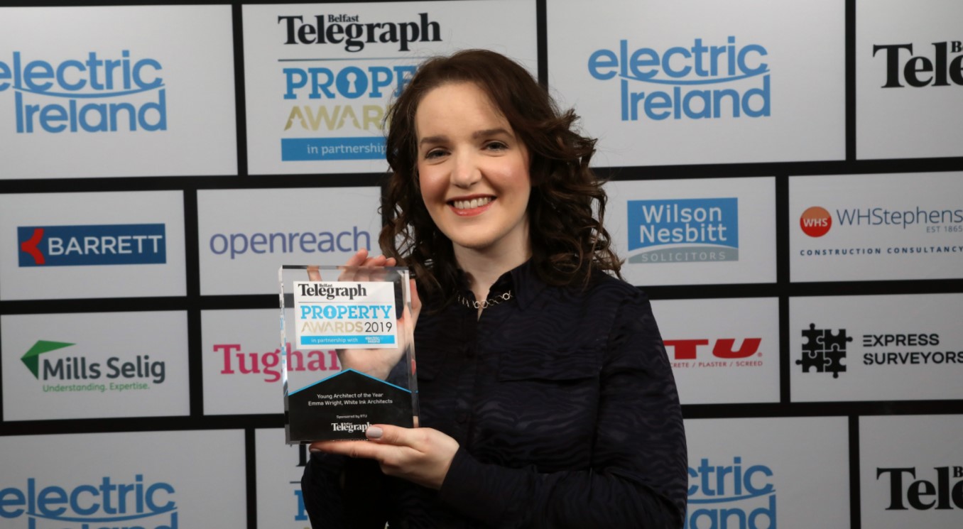 Emma Wright Young Architect of the Year with winner's trophy, standing in front of sponsors' board