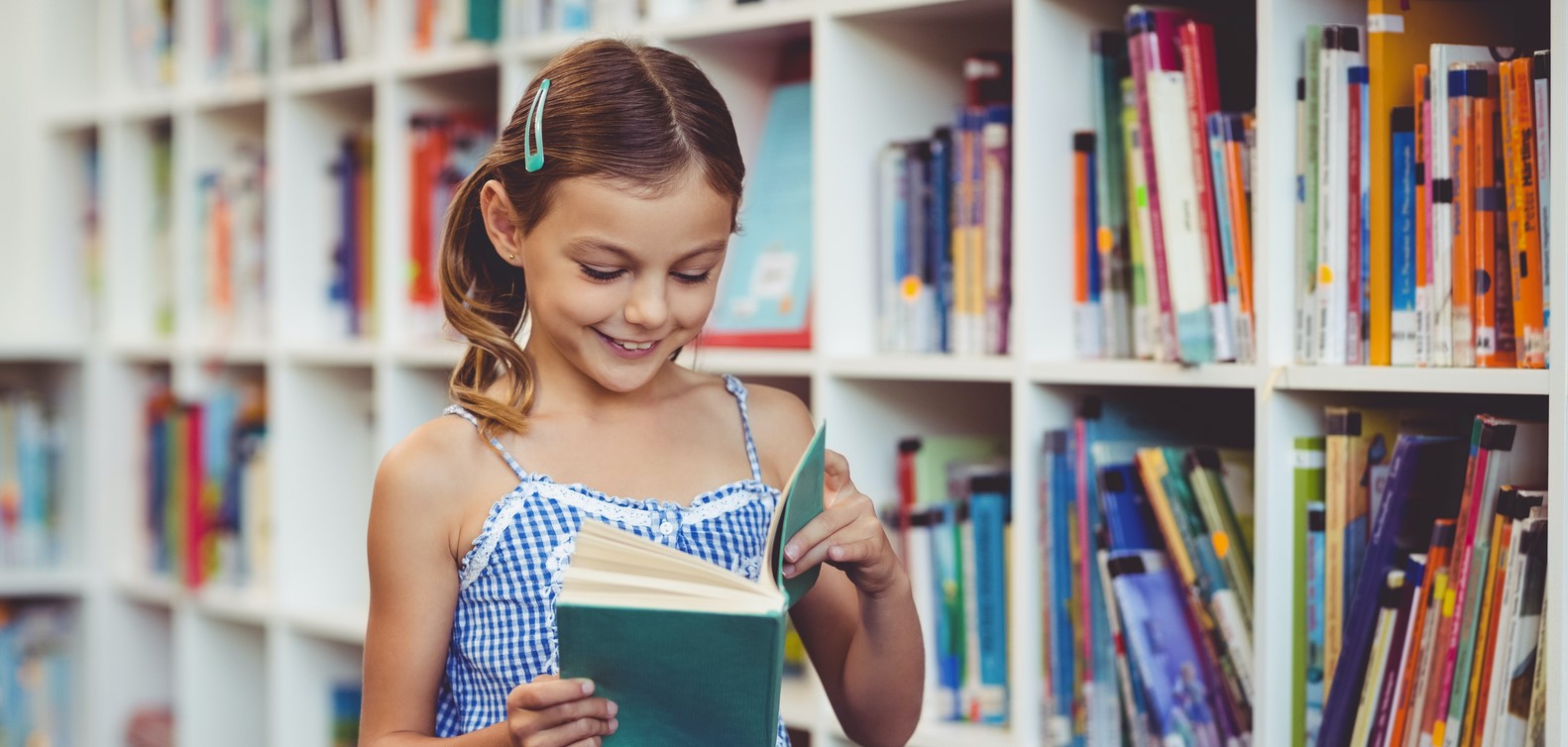 Smiling young girl in blue dress reading a book in a library