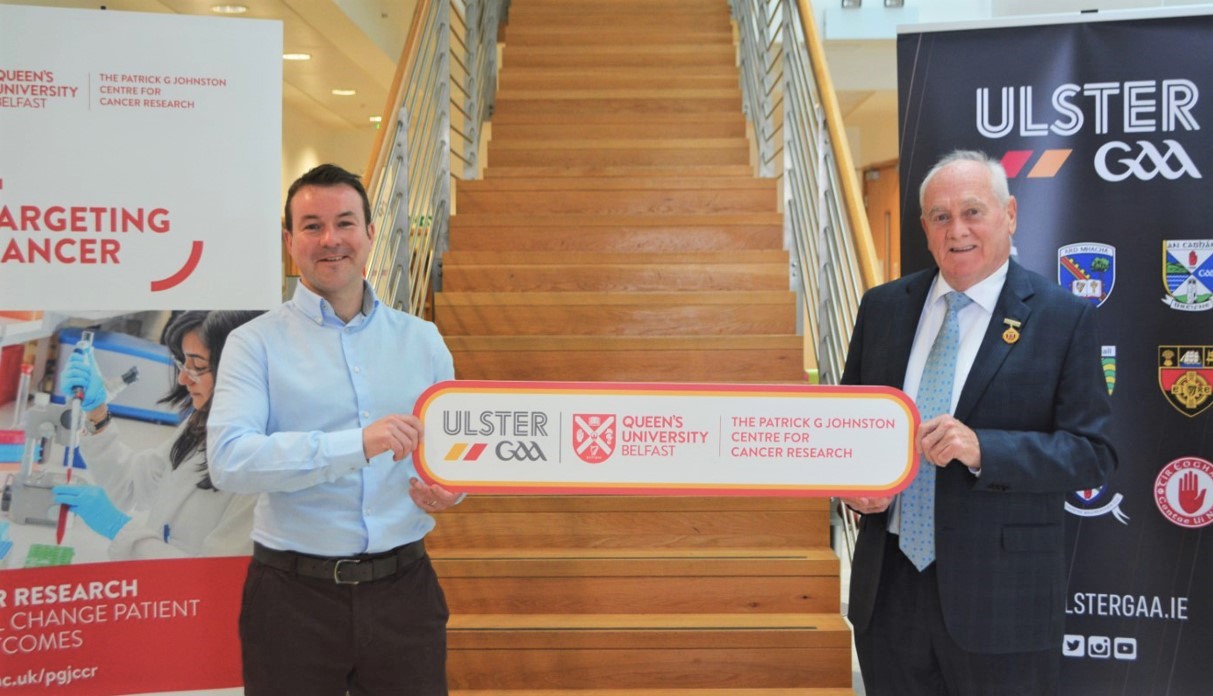 Dr Aidan Cole, Oncologist at Queen’s (standing left) with Oliver Galligan, President, Ulster GAA (right) at foot of stairs in Patrick G Johnston Centre for Cancer Research at Queen’s, holding banner  