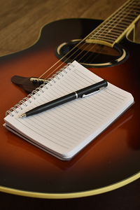 An acoustic guitar is sat on the floor with a blank lined notebook and a pen resting on top of the page.